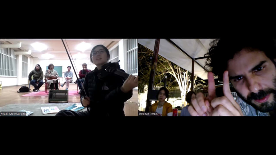 miaki holds the Cantini ready to play. stephan leans close to his camera and makes a gesture with both index fingers close to each other, pointed straight up.