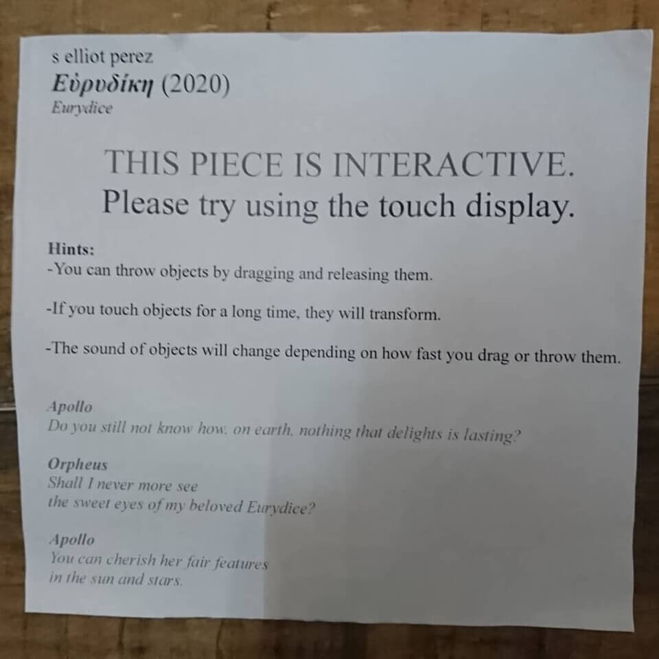 paper instruction sheet used at the exhibit. It says: 'THIS PIECE IS INTERACTIVE. Please try using the touch display. Hints: -You can throw objects by dragging and releasing them. -If you touch objects for a long time, they will transform. -The sound of objects will change depending on how fast you drag or throw them.'