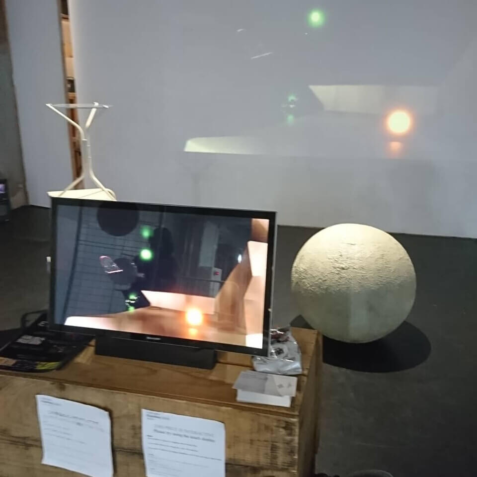 'Eurydice' is displayed on a touch monitor on a platform inside of the art gallery, BUoY. Behind the monitor is a stone sphere on the ground, about a meter high. On the wall behind it, the Eurydice feed is being projected.