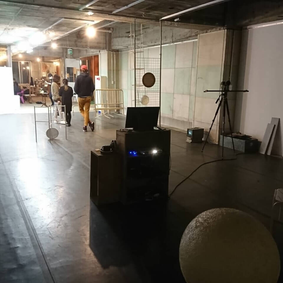 View from behind the monitor platform. The stone sphere is seen in the foreground. In the distance, three people are standing and conversing in front of the cafe space