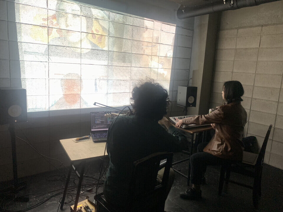 The performers, stephan e. perez and akari, sit, each on a chair in front of a small desk, facing away from the audience towards a bright wall projection. Stephan  is playing a MIDI violin with a bow and Akari is playing a MIDI keyboard with some knobs and levers. The projection shows an abstract, granular black and white pattern, blended with photos of people's faces and a bathroom tub.
