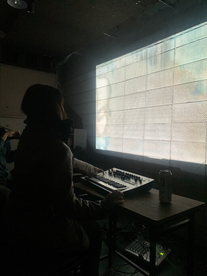In the darkened room, Akari pushes a small lever on the MIDI keyboard while looking at the bright projection. Part of a face can be seen on the left side of the abstract pattern.