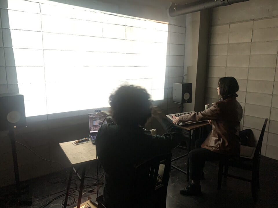 The performers sit, each on a chair in front of a small desk, facing away from the audience towards a bright wall projection. Stephan is playing a MIDI violin with a bow and a computer is in front of him. Akari is playing a MIDI keyboard with some knobs and levers. The projection is too bright to make out