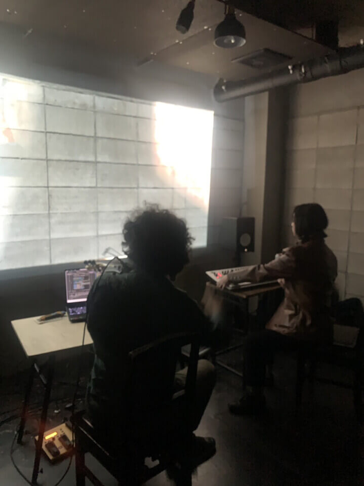 The performers sit, each on a chair in front of a small desk, facing away from the audience towards a bright wall projection. Stephan is playing a MIDI violin with a bow and a computer is in front of him. Akari is playing a MIDI keyboard with some knobs and levers. The projection is too bright to make out