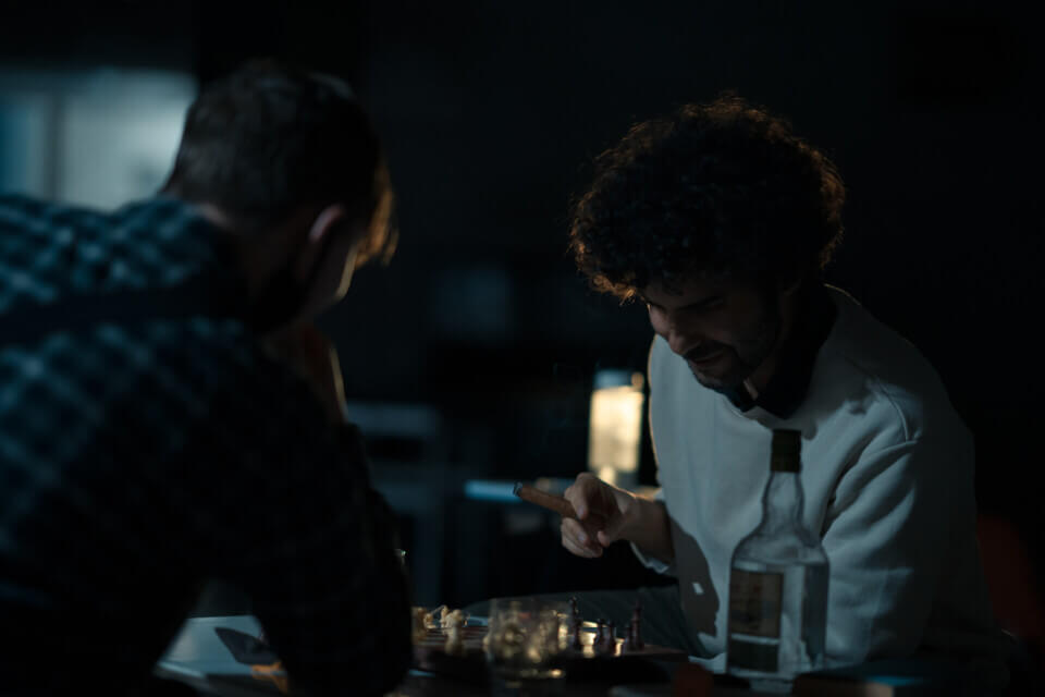 The performer plays sits at a larger table holding a cigar. A rum bottle is next to him and he looks down at a chessboard. Across from him, his opponent, in a plaid shirt, leans forward with interest.