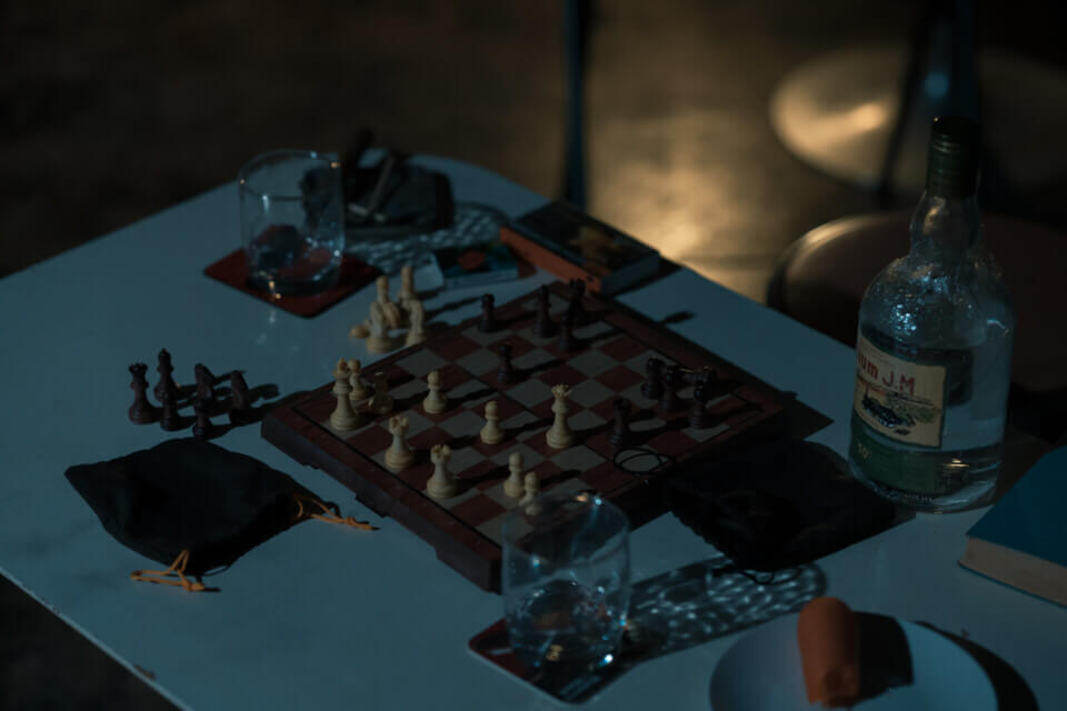 A chessboard showing an incomplete game, two glasses on coasters, a rum bottle, a book, a partly-eaten carrot and a cigar and matches in an ashtray on a long table.