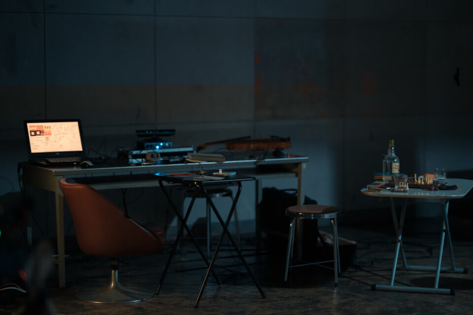 Full view of the stage. There's a large desk with a computer, a viola and other equipment, a small folding-desk, a larger folding-table with a chessboard and rum bottle on it and some chairs.