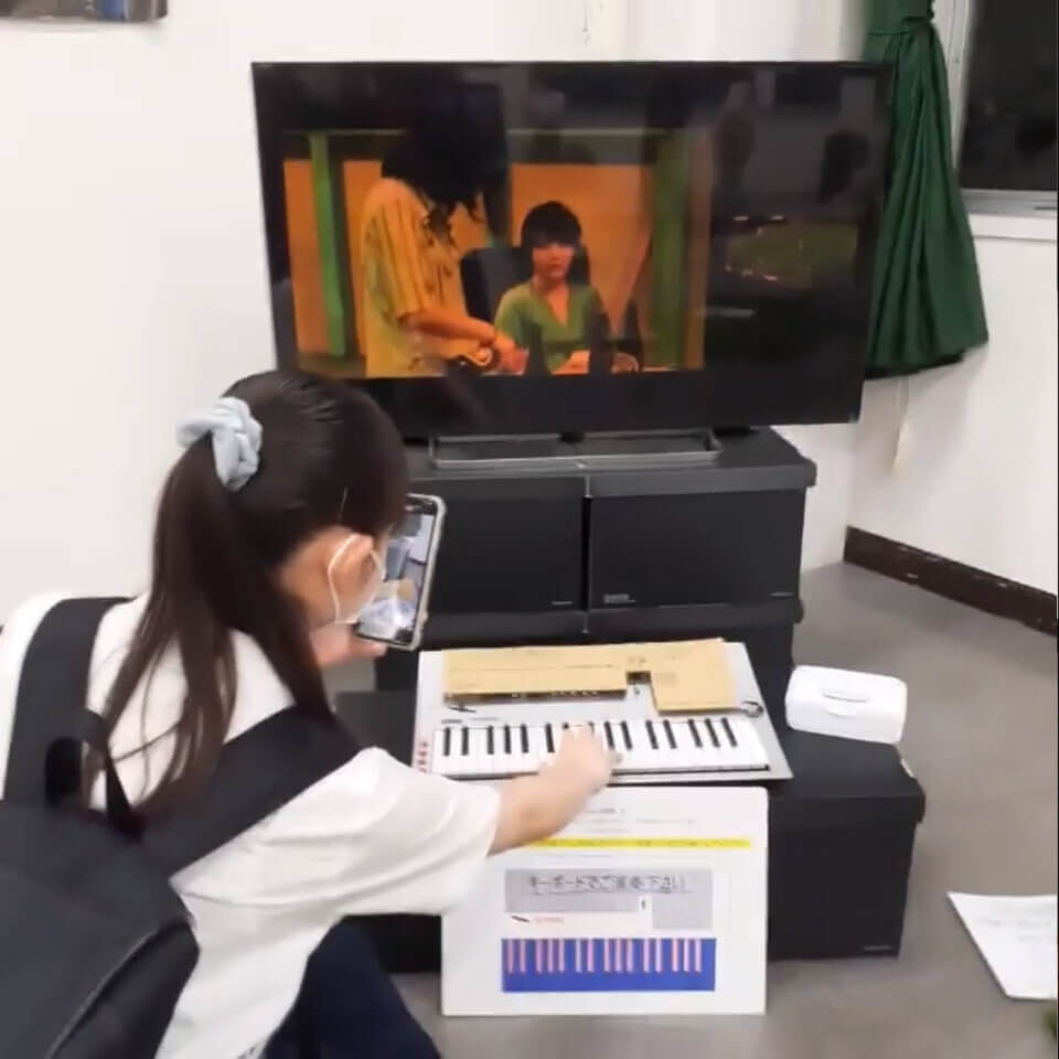 A young woman with a backpack plays the keyboard with one hand while videotaping herself with her phone. The monitor shows footage of a woman pouring beer for another woman.