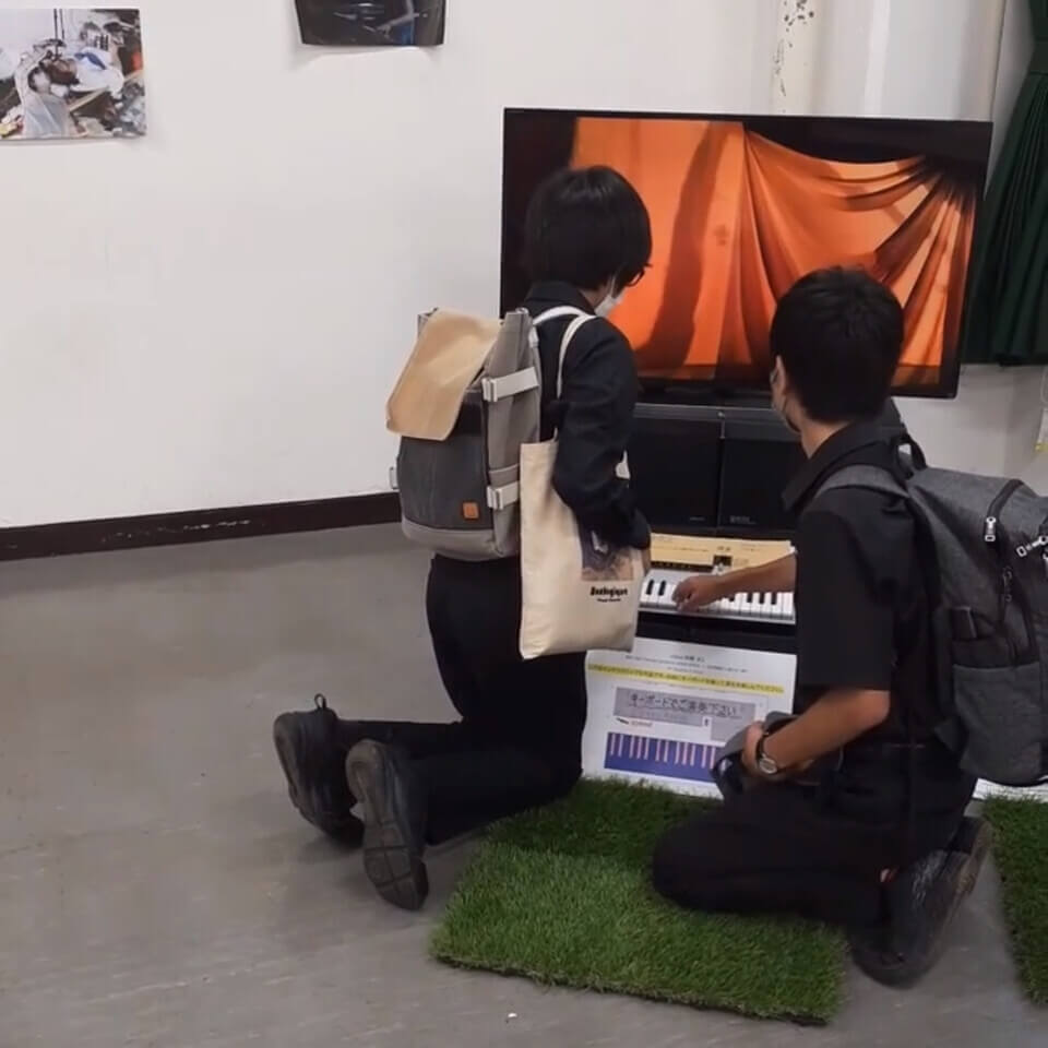 Two young men in black with backpacks crouch to play the keyboard. The monitor shows a red curtain.