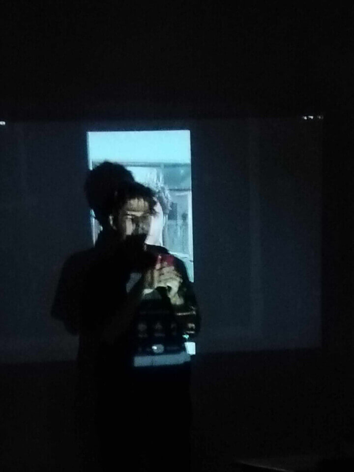 The performer, stephan e perez, does something on his smartphone while standing with his back close to a wall. A projection of a photo of himself illuminates him and the wall.