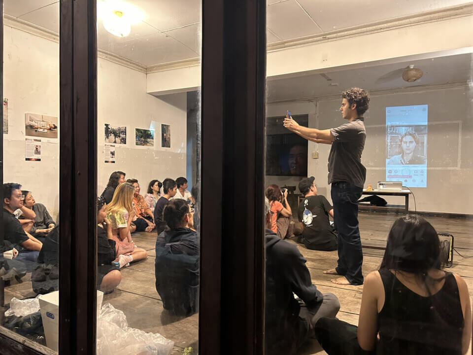 The performer takes a photo of himself in the lit room as the audience watches.