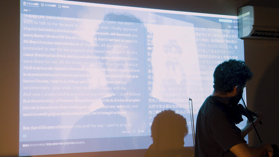 wall projection with an additive alpha composition consisting of several images. Recognizable elements are English and Japanese text from a screen capture, the performer's closeup, and a photo of a young woman. In front of the projection and two the right, the performer is playing a MIDI violin with a bow.