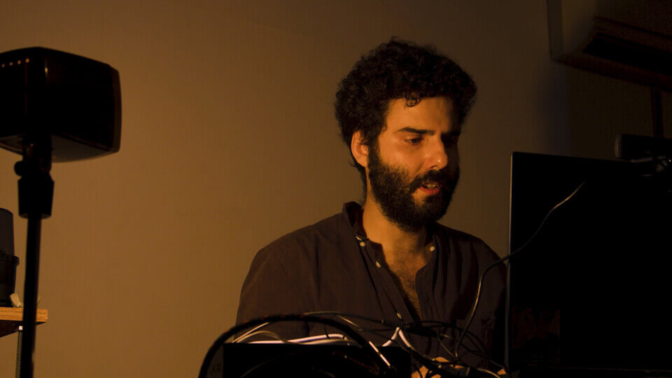 the performer sits in front of a laptop and types. Behind him, the wall is blank. His mouth is slightly open