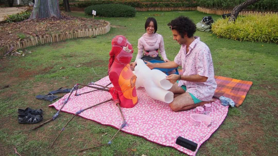 the artists, juno's jungle and stephan e perez, sit on the blankets of the play area. stephan slowly brings the plain white mannequin closer to the red spider mannequin