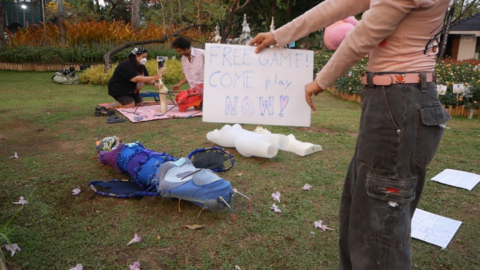 juno holds a sign up to the camera: free game! come play now! in the background, stephan and special guest mora are putting the mannequins in suggestion positions