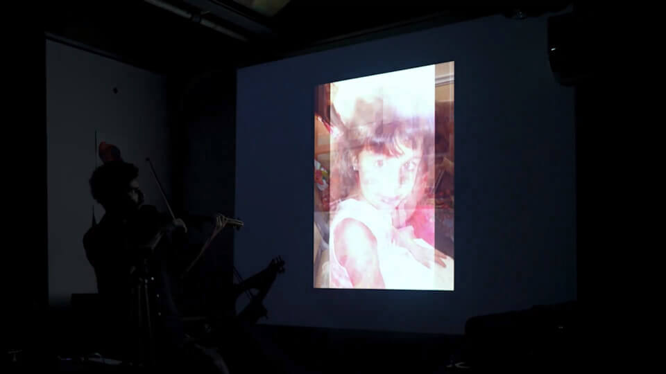 the performer, playing MIDI violin in front of a projection. The projection shows a composite of two images. One of the images is of a young girl, no older than 10, the performer's niece