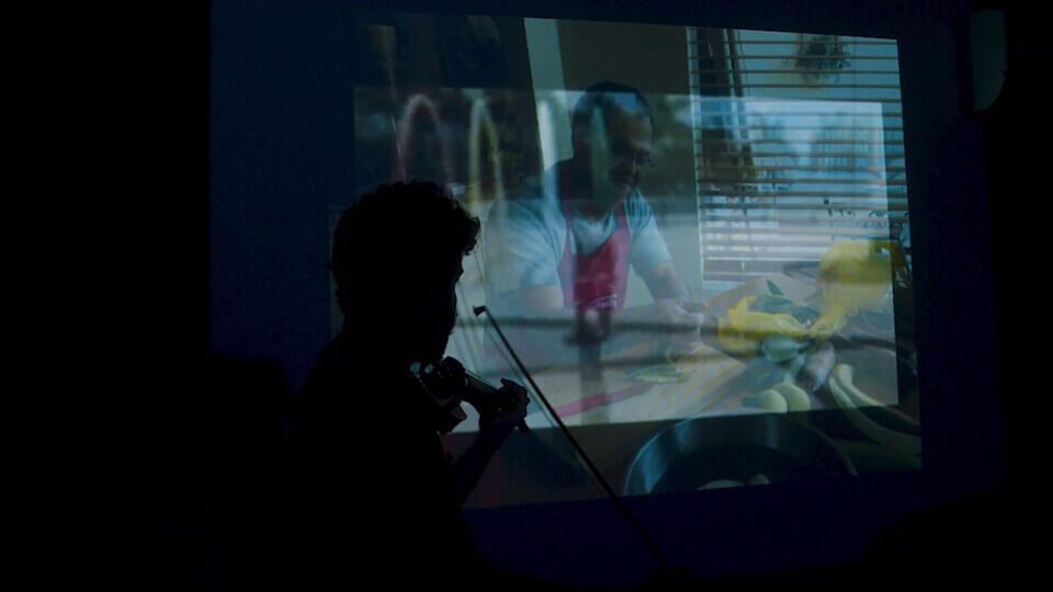 sillhouette of the performer, playing MIDI violin in front of a projection. The projection shows a composite of two images. One of the images is of the  performer's father. The other is of a pool area