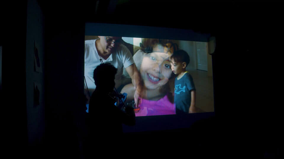 sillhouette of the performer, playing MIDI violin in front of a projection. The projection shows a composite of two images. One of the images is of the perforermer's father and a young boy, no older than 7, the performer's nephew. The other is of the niece.