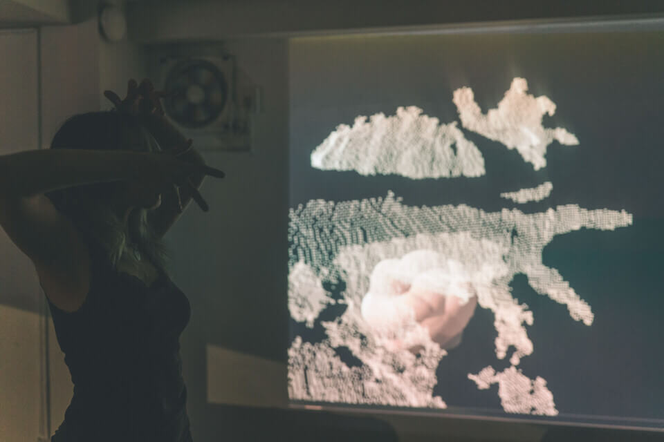 cat-o-nine-tails holds her hands in front of her face. Behind her, a wall projection shows a 3d cloud-point representation of her superimposed with a rounded, nucleus-like form