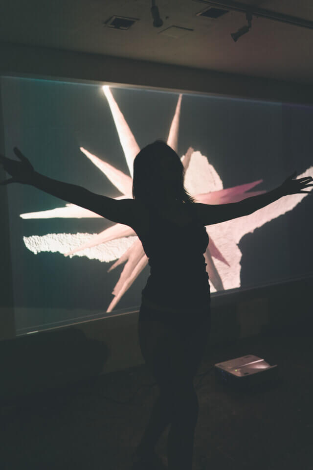 cat-o-nine-tails holds her arms to the side, she is visible as a sillhouette. Behind her, a wall projection shows a 3d cloud-point representation of her superimposed with a floating, 3d spiky form. The spikes are long and have a somewhat smooth texture