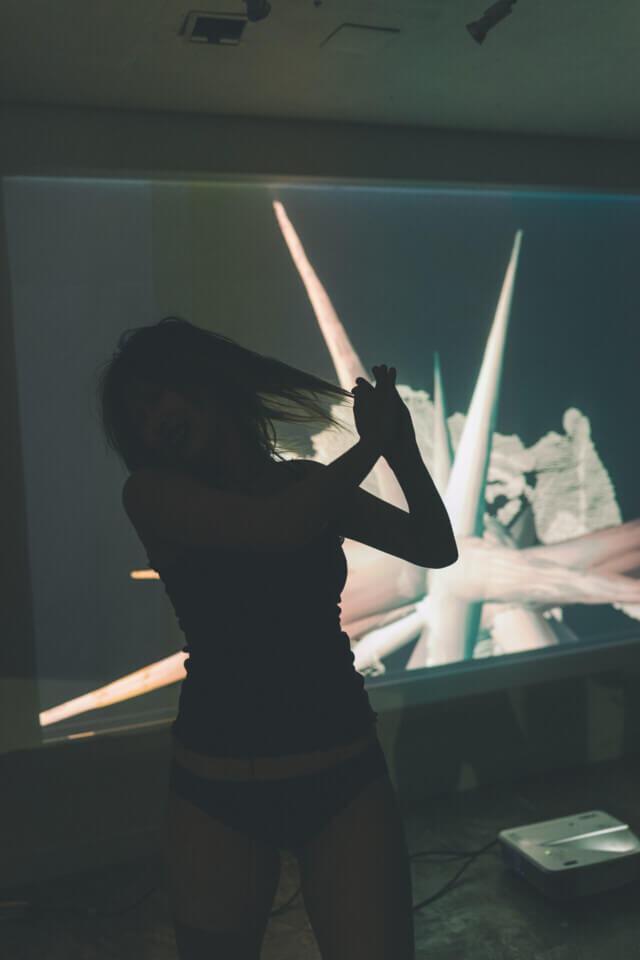 cat-o-nine-tails twists her head violently to the side and makes a blocking motion with her two hands in front of her. Behind her, a wall projection shows a 3d cloud-point representation of her superimposed with a floating, 3d spiky form. The spikes are long and have a somewhat smooth texture