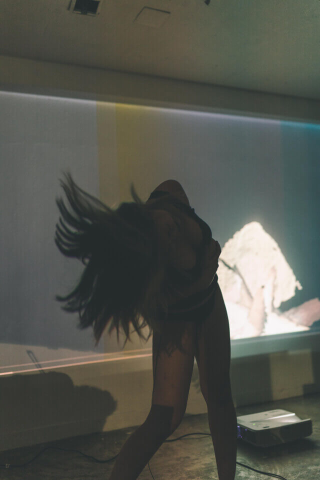 cat-o-nine-tails holds her sides and tosses her head downwards and to the side, her medium-length hair flying about. Behind her, a wall projection shows a 3d cloud-point representation of her