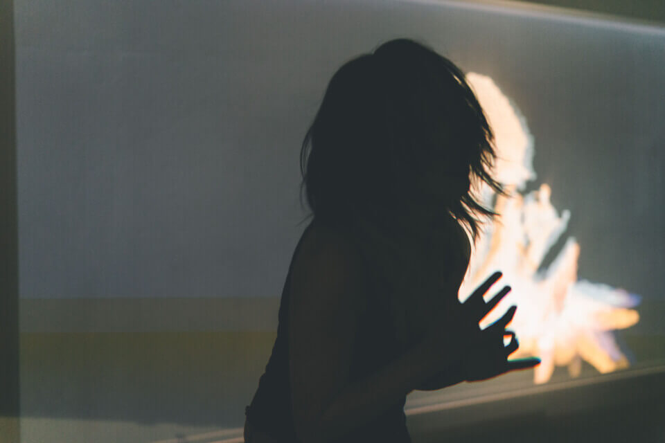 cat-o-nine-tails holds her hands in front of her and leans forward. Behind her, a wall projection shows a 3d cloud-point representation of her superimposed with a floating, 3d spiky form. The spikes are short and have a ripply texture