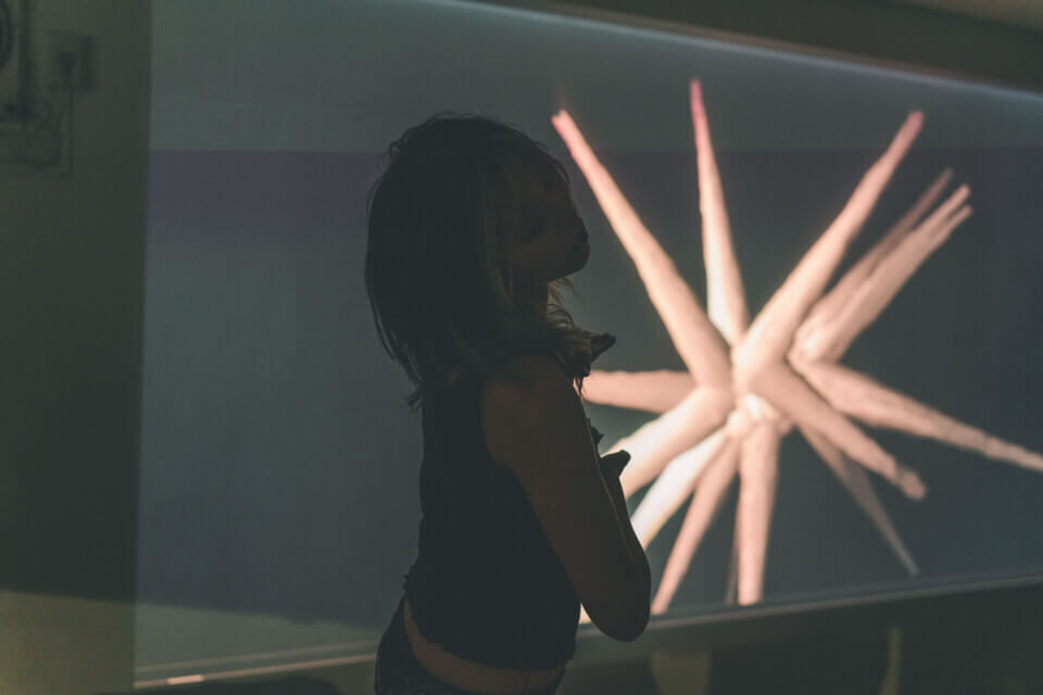 cat-o-nine-tails is in a dance-like pose with her head tilted, holding her hands with palms parallel, pointed to the side. Behind her, a wall projection shows a floating, 3d spiky form. The spikes are long and have a ripply texture
