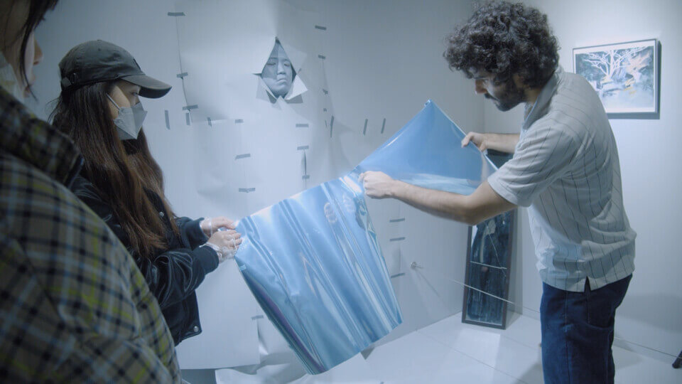 in a brightly lit area, a person helps hold a sheet of mirror paper as the performer cuts it in two