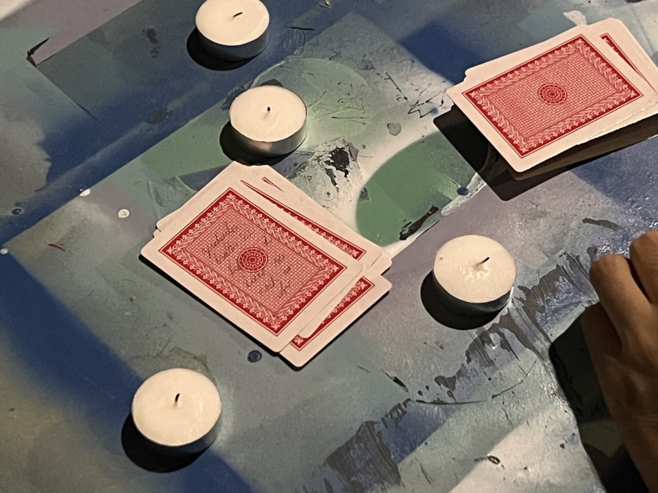 tea candles and playing cards on a table. The cards are stacked in two piles. The top card in the smaller pile has writing in pen that is hard to read