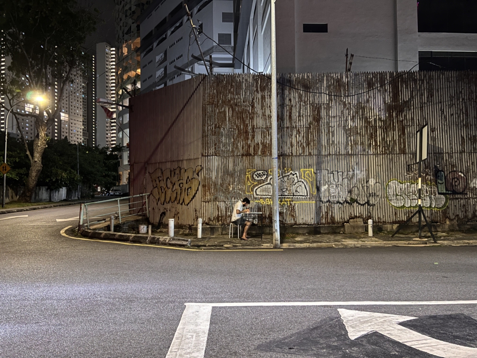 wider shot of the artist writing at the table. the surrounding road, trees and skyscrapers can be seen