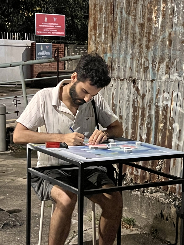 the artist writes on the back of a playing card at the table. Tea candles, cards and a jet lighter are in front of him