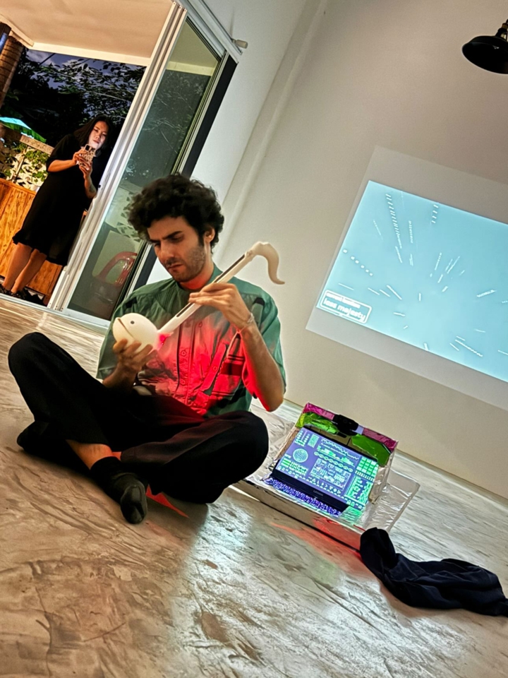 the artist is seated cross-legged, back to the console and viewport and playing an otamatone, an electronic instrument in the shape of a music note or tadpole. The viewport shows movement through space.