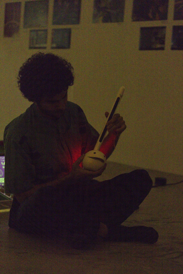 the artist is seated cross-legged in the dark, playing an otamatone, an electronic instrument in the shape of a music note or tadpole.