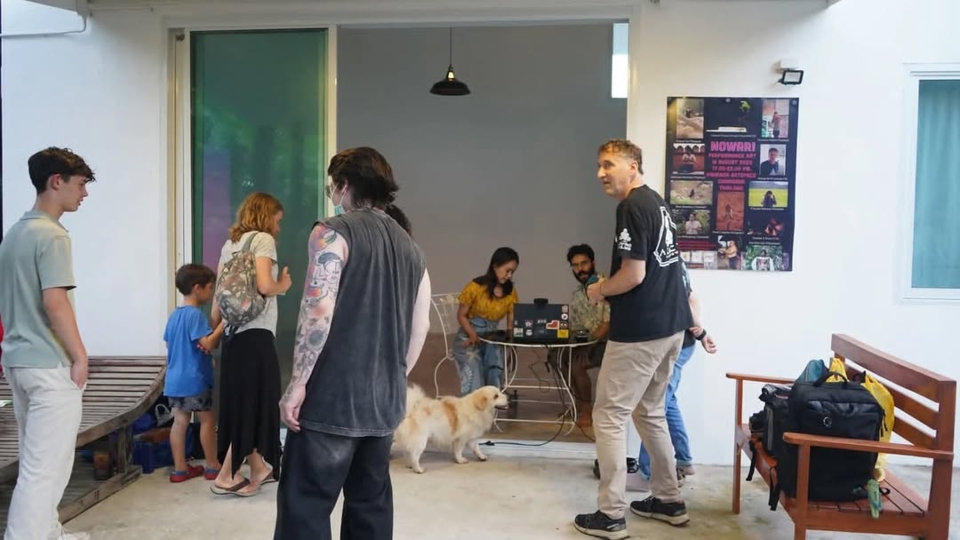 view of the artists seated at a small table inside the venue. View from outside, as the audience members walk towards the entrance.