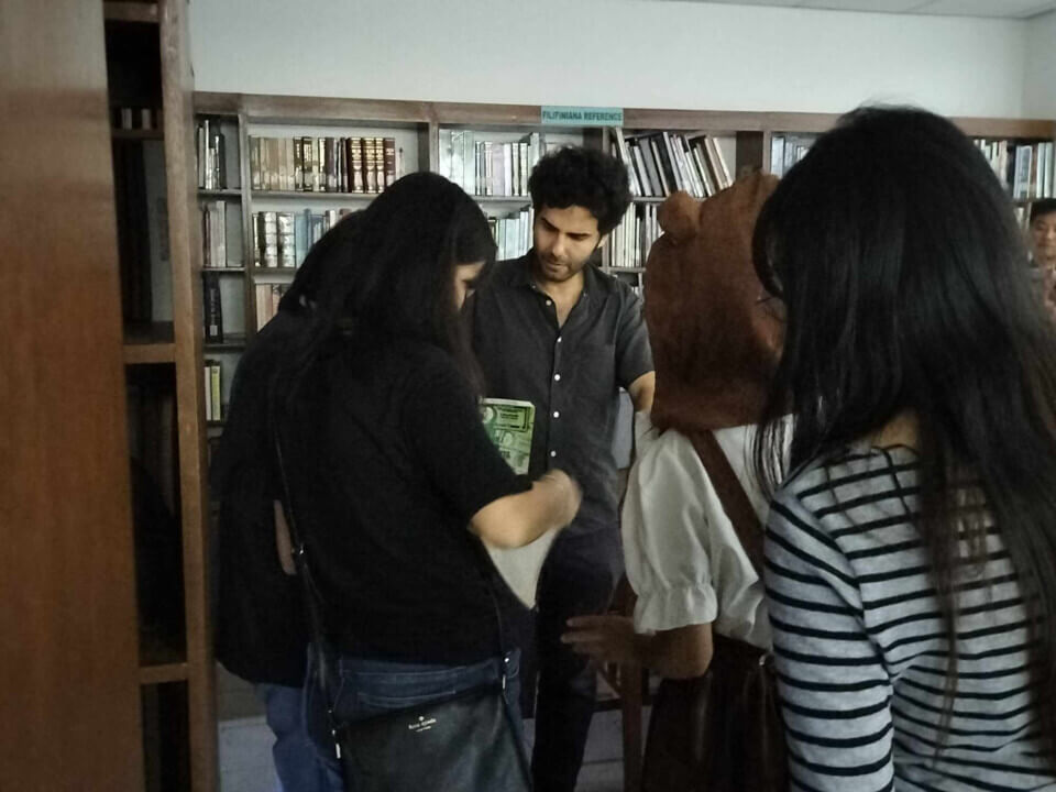 The performer, stephan e perez, stands in a library as group of students lines up to show him something