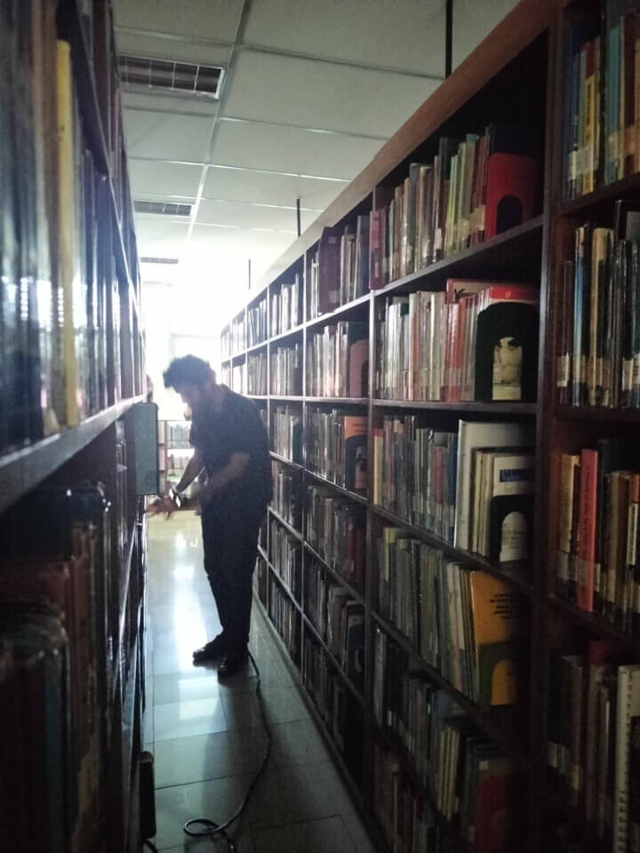 The performer stands alone in an aisle of bookcases, examing one of the shelves. A cable is coming from his feet