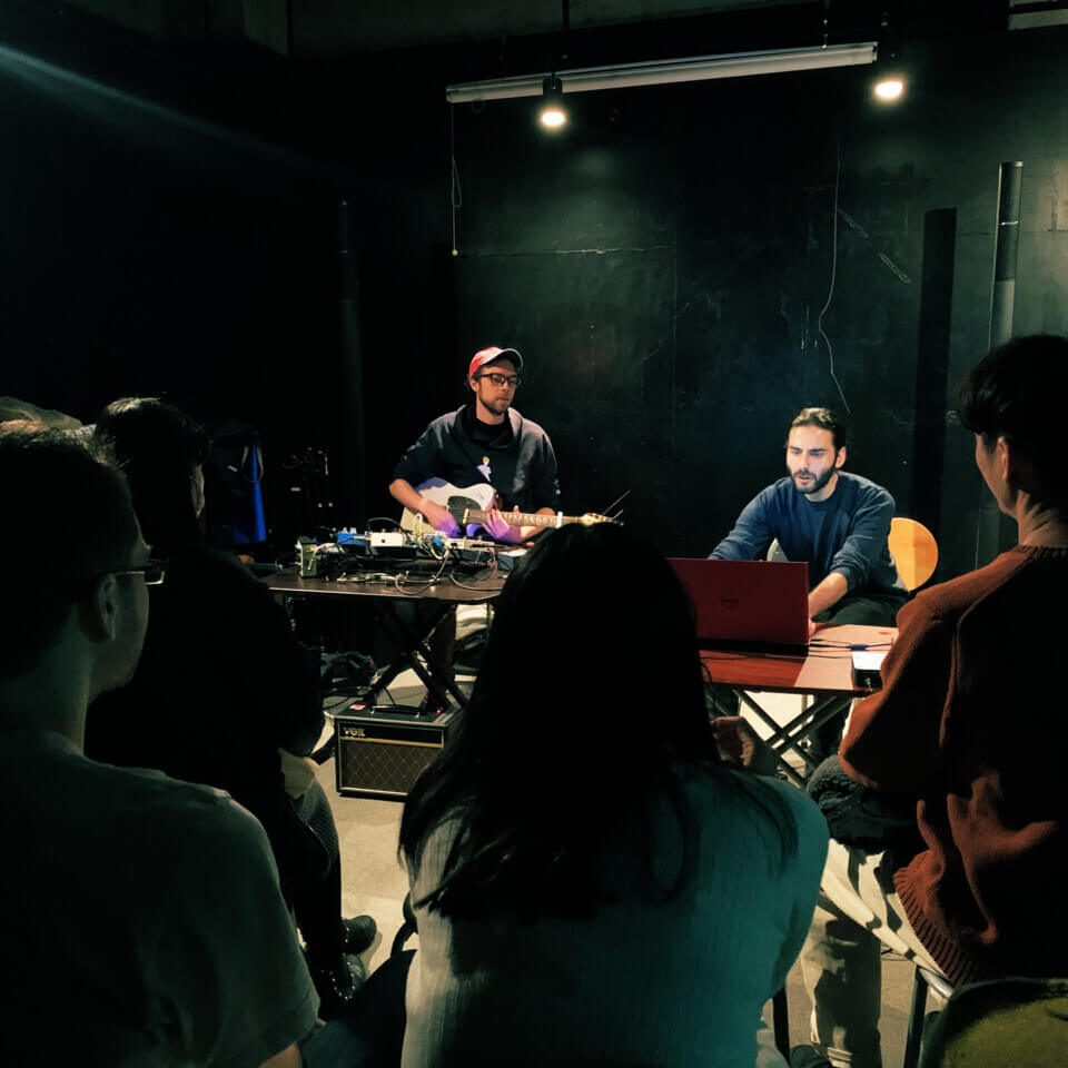 eric plays guitar and stephan does something on the computer. In the foreground, the audience is seated, listening