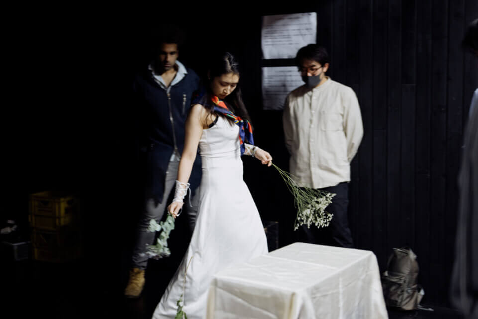 akari brandishes a bouquet of kasumi grass in each hand as she walks around a small table with a white tablecloth. She wears a white dress and colorful scarf. Two event guests stand behind her watching with their hands behind them.