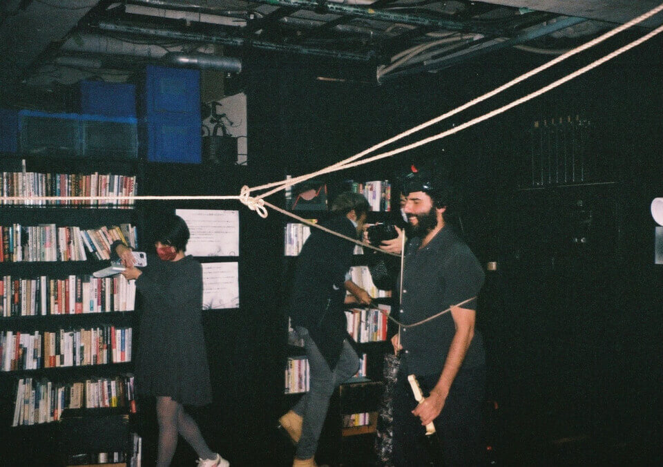 the rope is suspended across the room. one of the event guests is using one of its ends to tie up stephan, who laughs. another guest takes a photo with her phone