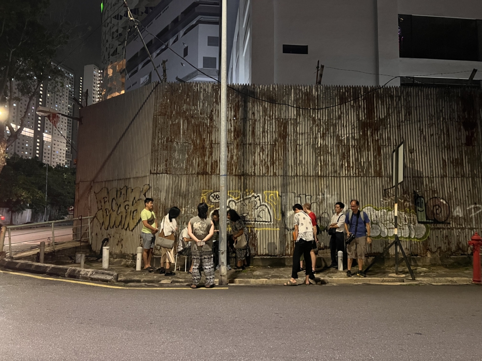 a group of about ten event visitors gathers around the artist, stephan e perez, as he writes at a table on the side of the road