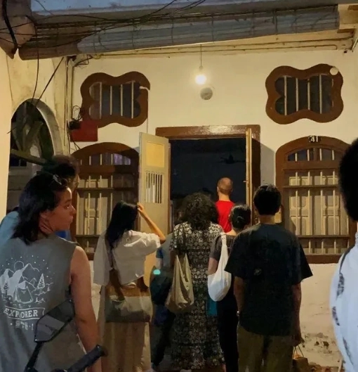 event visitors enter the newly opened double-doors to the residency house. The front of the house is lit by yellow lights but the inside is dark.