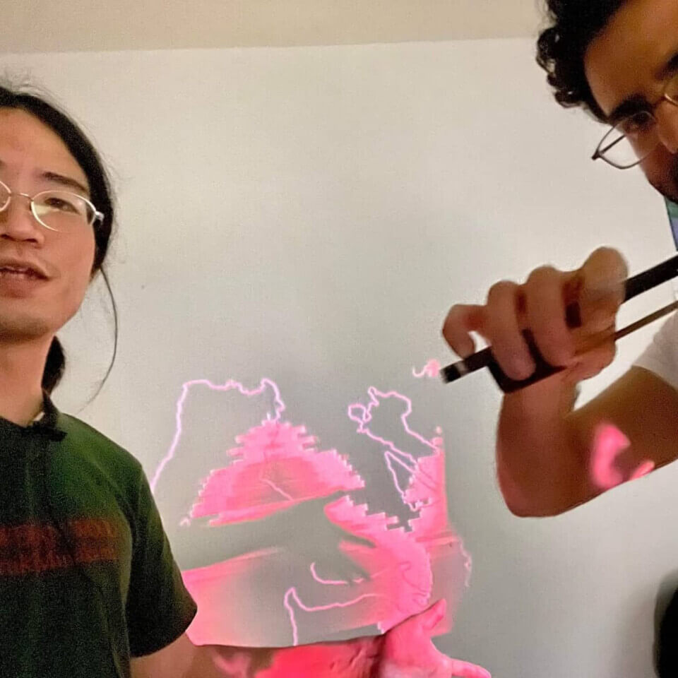 photo taken before the event of takumi-chan and stephan stand with the projection visible between him. takumi-chan is extending a hand in front of the projection and stephan is playing midi violin. The projection is of the red lines and curves used in their duo performance.