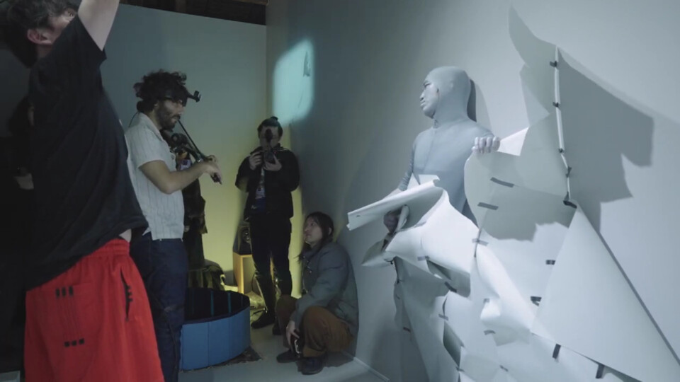 as event guests watch, takumi-chan, in his role as the wall man, slowly breaks out of the wall (wall-colored paper). stephan e perez plays the video violin, projecting a delayed image of takumi-chan onto the nearby wall.