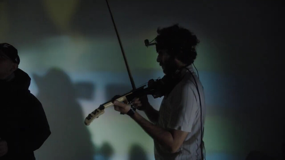 stephan e perez, a camera on his forehead, plays midi violin as unclear images and shadows fill the wall behind him