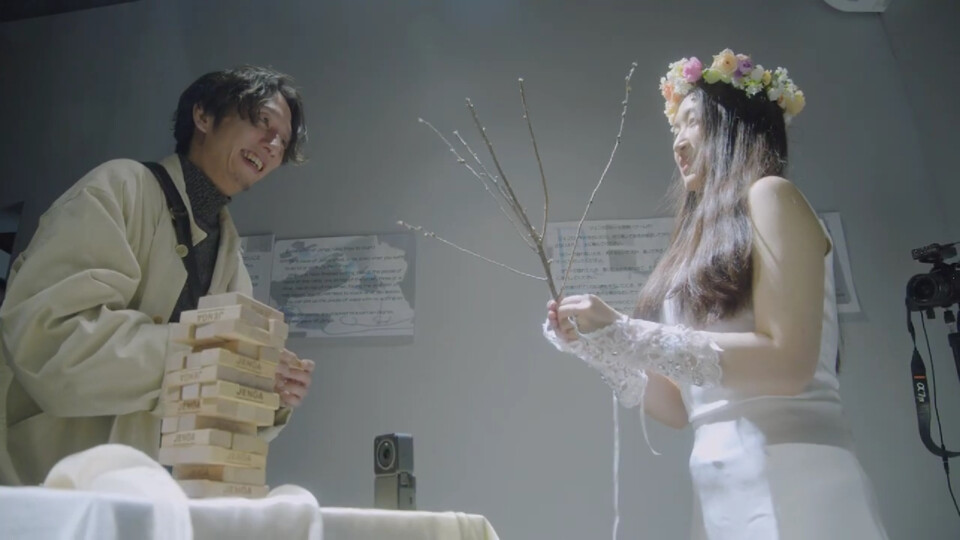 hagiwara akari stands holding a tree branch and smiles, facing an event guest holding a jenga block. In front of them, the jenga tower looms on the table