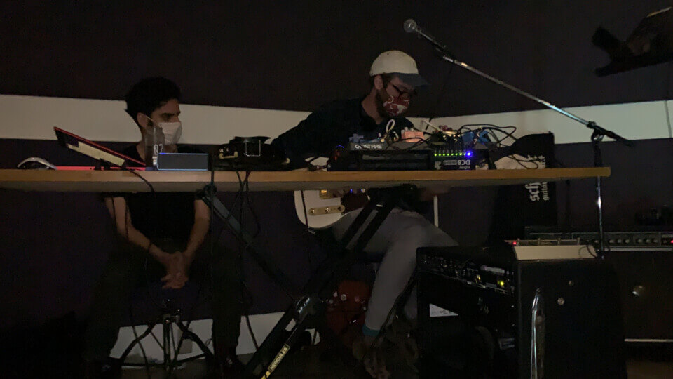 stephan, clasping his hands, sits and looks at eric, who is sitting next to him playing electric guitar.