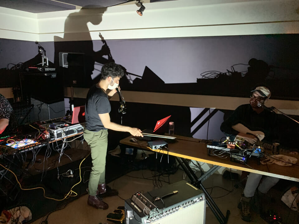 stephan places his bow on the table while holding the midi violin while eric beyeler sits at a table with effect pedals, holding an electric guitar