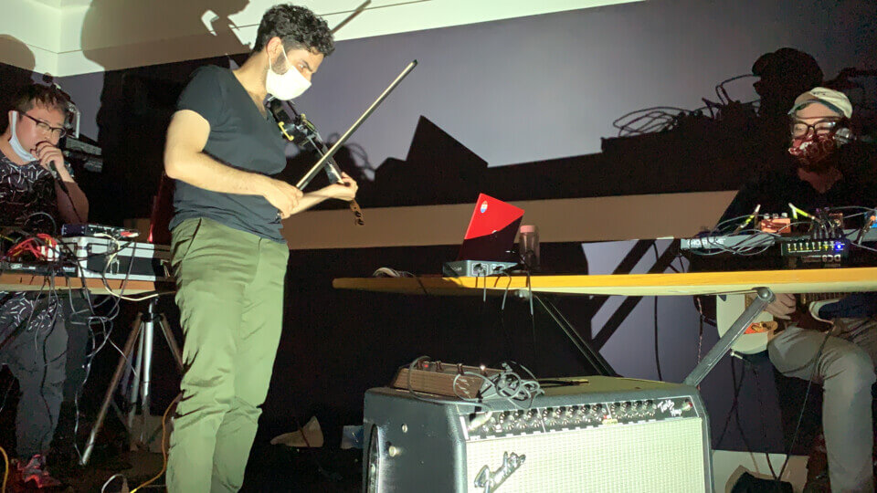 on the left, murakami hiroshi speaks into a microphone. stephan e perez holds his midi violin while eric beyeler sits at a table with effect pedals, holding an electric guitar